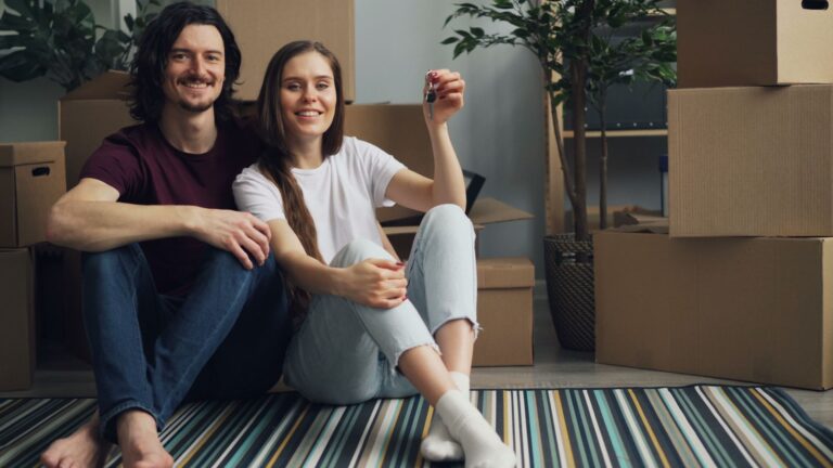 A joyful couple sitting on a striped carpet with moving boxes, holding a key to their new home.